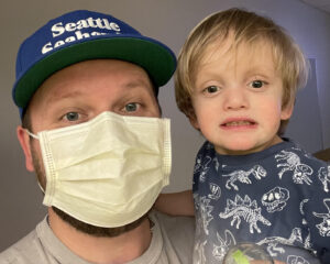 A father wearing a blue baseball cap and face mask holds up his son for a photo. The boy has blond hair and is wearing a navy dinosaur shirt.