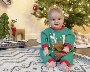 An 11-month-old boy sits in front of a lit-up Christmas tree. He's wearing a red and green onesie with a reindeer on the front and is smiling at the camera. There are various ornaments on the tree and other Christmas decorations in the background.