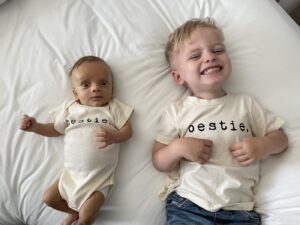 Two brothers, one 3 months old and one 3 years old, lie on their backs on top of a white comforter. The baby is wearing a onesie, while the toddler is wearing a T-shirt, but both pieces of clothing are cream-colored and say "bestie" in dark writing. The baby's eyes are wide open and looking at the camera, and the boy is smiling with his eyes shut. 