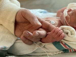 A small, premature baby boy lies on a blanket in a bed in the NICU. He's hooked up to various tubes and monitors and is holding his dad's pointer finger in his left hand.