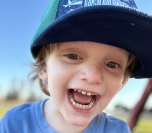 A young boy smiles broadly while wearing a baseball cap and blue T-shirt. We can tell he's standing outside on a sunny day, though the background is blurred.