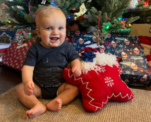 A healthy baby sits smiling in front of a Christmas tree, with a stocking and some gifts next to him. 