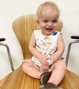 A 3-year-old boy sits in a large, wooden chair in a nondescript doctor's office with white walls. He's sharing a big smile, and two tiny bottom teeth are visible . He crosses his hands on his lap and has a medical wristband on his left wrist.