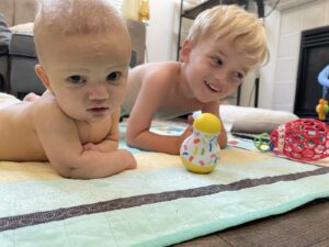 Two small boys lie shirtless on their stomachs on a baby mat, with toys scattered in front of them. The baby stares directly into the camera, while the older brother smiles mischievously and looks off to the right of the frame.