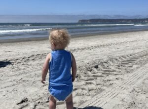 A toddler in a blue onesie stands on a sandy beach and looks out at the ocean during a sunny day.