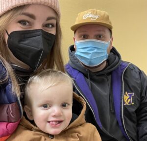 A wife, husband, and 3-year-old boy take a selfie in a room with yellow walls. The mother and father are wearing medical masks, while the boy sticks his tongue out for the camera. 
