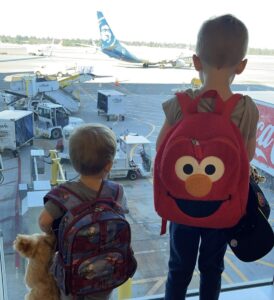 Two young boys - about the ages of 3 and 5 - stand in front of a large window at an airport looking down on the planes and airport workers below, at an airport gate. We see the backs of the children, and the boy on the right is wearing a large, red Elmo backpack. The smaller boy on the left also is wearing a backpack and carries a large teddy bear. 