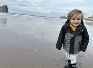A 3-year-old grins as he stands on a beach at low tide. He's wearing a big jacket and rain boots. 