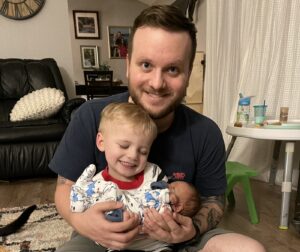 A young father holds his two boys - one is a bit older than a toddler and the other is a newborn. Both are held in the father's lap as he smiles at the camera. They're sitting in a living room.