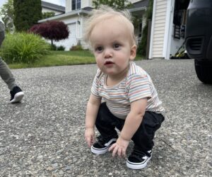 An 18-month-old baby squats down on the driveway of a home, looking just to the left of the camera. He's wearing a tiny pair of black sneakers. 