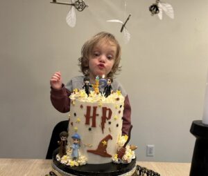 A young boy with long blond hair faces a large white birthday cake, which has the dark red initials "HP" on the front, for "Harry Potter." The cake features decorations like a sorting hat and Harry Potter characters, and a candle appears to be on the top.