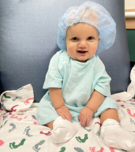An infant is pictured in a blue mesh cap, pale green hospital gown, and white socks. He sits with his back to a gray curtain and on a white bedspread with multicolored footprints.