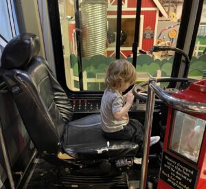A young boy with longish blond hair in a gray shirt sits in a large black chair before a steering wheel. 