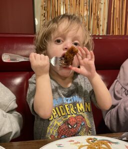 A toddler wearing a Spider-Man T-shirt holds a fork awkwardly and shoves a piece of meat into his mouth using both hands. 