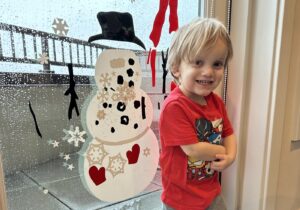 A 3-year-old stands in front of a stylish piece of art portraying a snowman with a large black hat. The boy smiles proudly, arms folded in accomplishment.