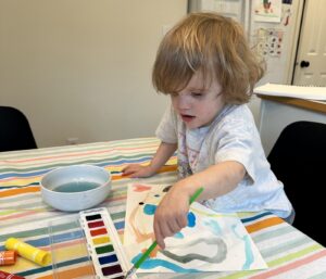 A 3-year-old boy sits at a table painting with watercolors. He's holding a brush in his left hand and dipping it into a dark paint color, and he looks focused. 