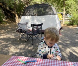 A toddler in pajamas with stars fiddles with something while sitting at an outdoor picnic table in the woods. A tent is set up behind him.