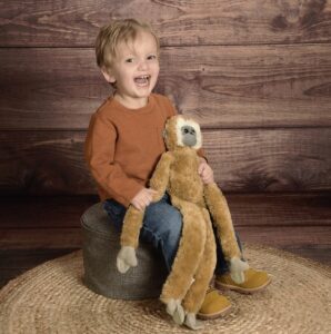 A young boy sits on a shot stool on a brown rug, with his back to a darker brown wooden wall. He wears a brown shirt and jeans and holds a large, rather flat toy resembling a monkey.