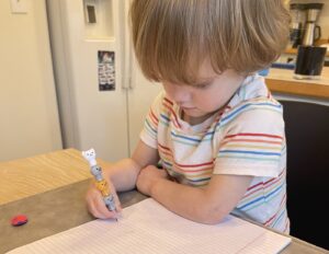 A young boy with long, thick, brownish-blond hair draws on what appears to be a notepad on a table with different shades of brown. He wears a white shirt with red, orange, yellow, green, and blue stripes.