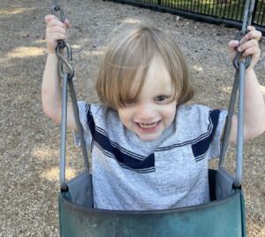 A young boy sits in a swing, holding on to the chains and smiling at the camera.