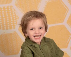 A young boy with dark blond hair in a forest green shirt smiles in front of a yellow wall with geometric shapes.