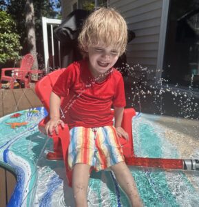 A young boy, maybe 3 or 4 years old, laughs while sitting in a small red chair atop a shallow inflatable pool. The edges of the pool are spraying water up at the boy, who's wearing a red swim shirt and striped swim trunks.