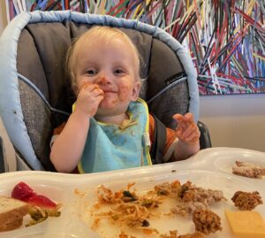 A smiling toddler with food smeared all over his face sits in a baby chair with food all over it. 