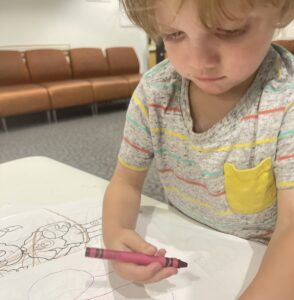 A young boy in a gray shirt with colorful stripes holds a magenta crayon as he considers a coloring page. He appears to be standing at a low table in a waiting room.