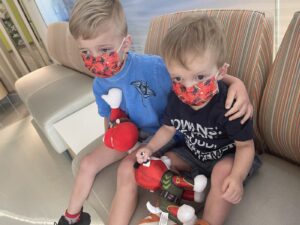 Two young boys sit on a couch in a waiting room at a hospital. Both are wearing red face masks and holding toys in their laps. The older boy has his left arm around his younger brother's shoulder.