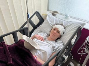 A woman with her forehead bandaged lies in a hospital bed. She's in a hospital gown, with a white sheet and a maroon blanket over her. A white hat sits on her midsection.