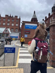 A father with a backpack carries his 3-year-old daughter on his shoulders as they walk toward a U.K. hospital. We see the two from behind, and the young girl is wearing a baseball cap. The sky is cloudy and gray.