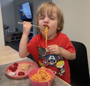 A young boy closes his eyes and smiles happily while taking a big bite of spaghetti. He's wearing a red T-shirt and looks to be sitting at a table, with a TV in the background in the next room. 