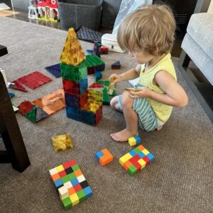 A blond boy kneels next to a small structure he's built with colorful magnet tiles. There are blocks and toys strewn around him, and he looks to be at home, perhaps in a living room or playroom. 