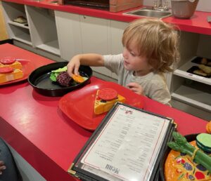 A young boy stands at a counter in a play kitchen with red countertops. Several plates with plastic foods are in front of him, along with a menu. He appears to be placing something on top of a burger patty.