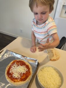 A young boy, about 3 years old, stands on a chair next to the kitchen counter. In front of him is a small unbaked pizza on a baking sheet and a bowl of shredded cheese. He's putting the cheese on the pizza and appears focused on the task at hand.