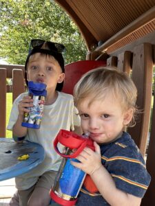 Two young boys smirk at the camera while drinking from water bottles. The younger boy, seated closer to the camera, is wearing a striped shirt and holding a red bottle with both hands. The older boy, seated just behind him, has on a hat and sunglasses resting atop his head, and he's holding a blue bottle with his right hand. The boys appear to be in some sort of outdoor wooden play structure; grass and a large tree are visible behind them.