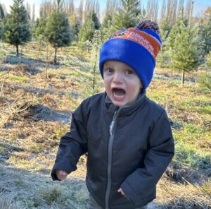 A young boy with a wide open mouth stands outside at a tree farm. He's in a dark gray jacket and a red and blue knit hat, and he's clenching his arms.