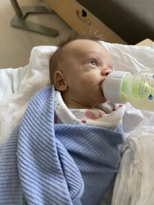 An infant boy is wrapped in a blue blanket and lies on white bedding. His head is turned to the left, and he's sucking on a bottle of formula.