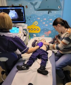 A 3-year-old girl lies prone on a hospital bed as her mother sits to her right and holds her while a medical worker conducts an ultrasound scan on her stomach. The medical worker is looking at the screen to the left and the mother is looking down at her child. 