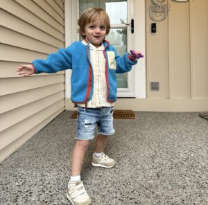 A boy is outside the front door of a house painted in a light tan or beige. He's blond and wears tennis shoes, blue jean shorts, a white shirt, and a bright blue sweater with some sort of emblem on one side.