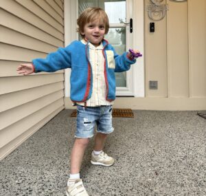 A young blond-haired boy stands outside the doorway of a tan house. He wears a white shirt, blue jean shorts, white tennis shoes, and a bright blue jacket.