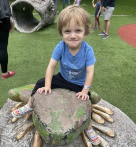 A blond boy sits at what appears to be a gray and green bench, his hands in front of him on what seems to be a gray and green stone, looking something like a crocodile or alligator head. The boy wears a blue T-shirt and dark pants with multicolored shoes; behind him appears to be artificial turf with playground items, with the legs of others present.