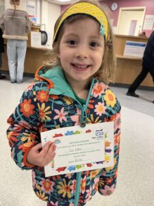 A 4-year-old girl with a colorful flowered jacket accented by a bright yellow headband stands in a hospital lobby holding a colorful certificate of bravery. She's smiling proudly.