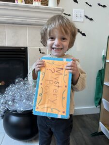 A 3-year-old boy with bushy, blond hair smiles from ear to ear as he holds a yellow and blue box marked "Mystery Box." He's standing in front of a white fireplace in a living room.