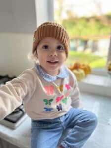  A young girl in a brown cap, a tan sweater with colorful butterflies, and blue jeans. She is sitting on a countertop with a window in the background.