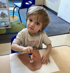 A blond-haired young boy sits at a desk with what appears to be a glue stick over some brown paper.