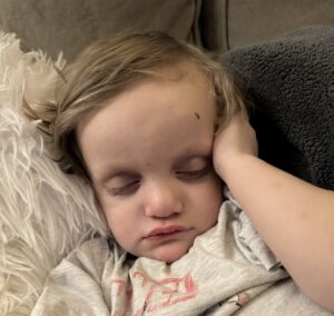 A young blond-haired boy is wearing a light gray shirt with a red design while his head rests on a white pillow. His eyes are closed.