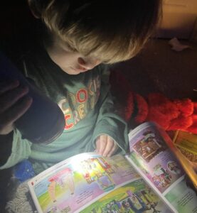 A 3-year-old boy sitting in bed uses a flashlight to look at a children's book. The light from the flashlight and the shadows from the dark room have an interesting interplay. 