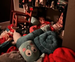 A massive pile of stuffed animals thrown on the floor. A young boy's face is barely visible in the middle of the pile. 