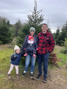 A family of four poses for a photo in front of a small Christmas tree at a tree farm. A man and woman are standing front and center, and the woman has a baby strapped to her front. A young girl about 4 years old stands to the woman's side, holding her hand. All are bundled up in warm clothing. There's no snow visible, but it appears to be a cold and cloudy day.